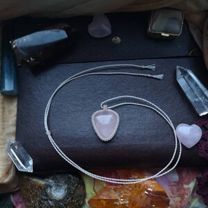 May include: A pink heart-shaped crystal pendant on a white cord necklace. The necklace is lying on a brown leather surface with other crystals and stones.