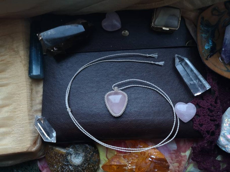 May include: A pink heart-shaped crystal pendant on a white cord necklace. The necklace is lying on a brown leather surface with other crystals and stones.