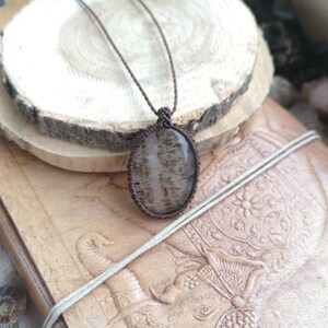 May include: A brown leather cord necklace with a light brown and gray agate pendant. The pendant is a natural stone with a unique pattern.