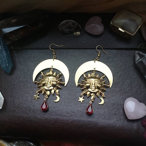 May include: Gold tone earrings with a crescent moon and sun face design. Each earring has a dangling red teardrop crystal.