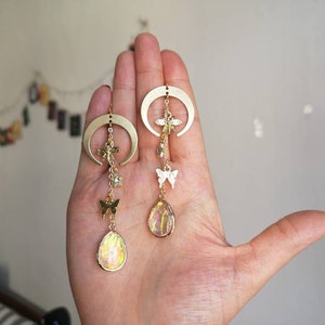 Puede incluir: Un par de pendientes de media luna dorados con colgantes. Cada pendiente tiene una piedra iridiscente en forma de l&aacute;grima, un colgante de mariposa y un colgante de lib&eacute;lula.