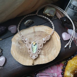 Puede incluir: Un delicado collar de macram&eacute; con un cord&oacute;n marr&oacute;n claro y un colgante de labradorita verde claro. El collar se muestra sobre una superficie de madera con otros cristales.