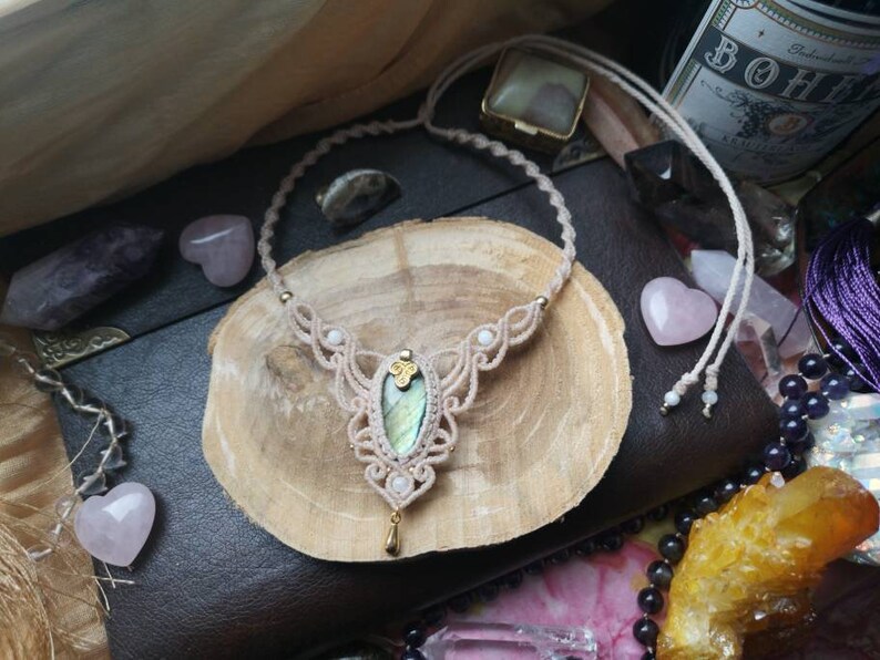 Puede incluir: Un delicado collar de macram&eacute; con un cord&oacute;n marr&oacute;n claro y un colgante de labradorita verde claro. El collar se muestra sobre una superficie de madera con otros cristales.
