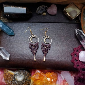 May include: A pair of maroon and gold earrings with a crescent moon charm and a macrame design.