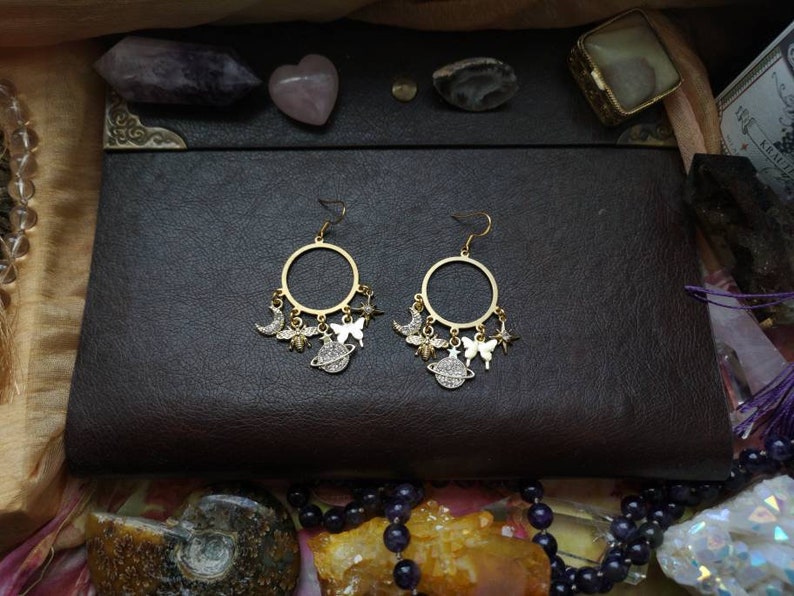 May include: Gold hoop earrings with dangling charms, including a crescent moon, a butterfly, a planet, and a star. The charms are silver with clear rhinestones.