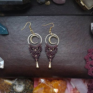 May include: A pair of maroon and gold earrings with a crescent moon charm. The earrings are made with macrame and have a beaded design.