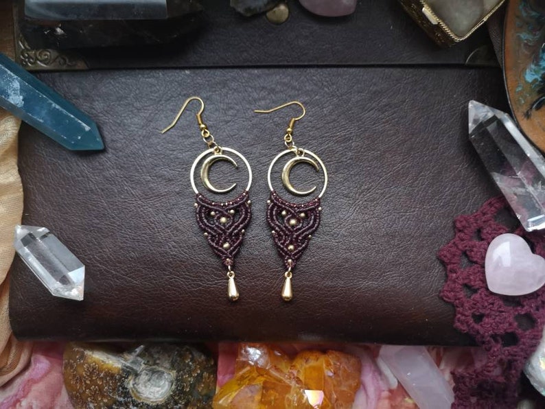 May include: A pair of maroon and gold earrings with a crescent moon charm. The earrings are made with macrame and have a beaded design.