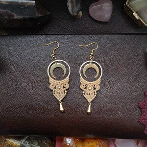 May include: A pair of gold earrings with a crescent moon charm and a delicate macrame design in a beige color.