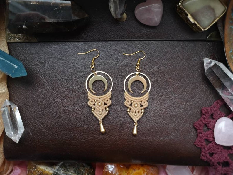 May include: A pair of gold earrings with a crescent moon charm and a delicate macrame design in a beige color.