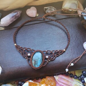 May include: A brown leather cord necklace with a blue and green gemstone pendant. The pendant is surrounded by smaller white gemstones and gold beads. The necklace is on a brown leather surface.