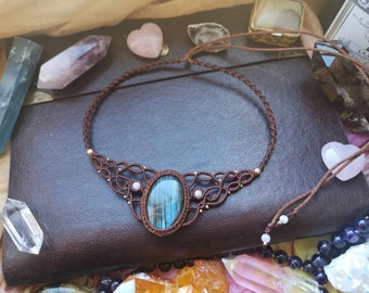 Puede incluir: Un collar de cordón de cuero marrón con un colgante de piedra preciosa azul y verde. El colgante está rodeado de piedras preciosas blancas más pequeñas y cuentas doradas. El collar está sobre una superficie de cuero marrón.