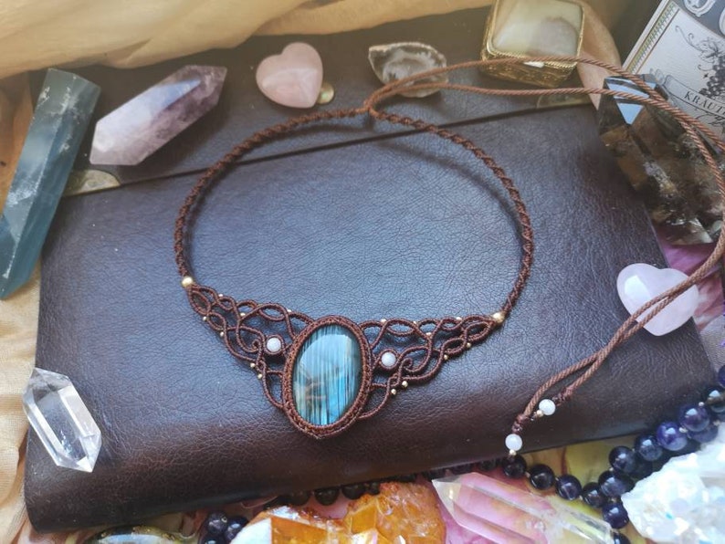 May include: A brown leather cord necklace with a blue and green gemstone pendant. The pendant is surrounded by smaller white gemstones and gold beads. The necklace is on a brown leather surface.