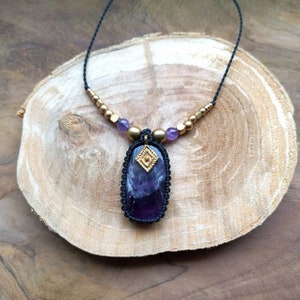 May include: A purple amethyst gemstone pendant necklace with gold accents and black cord. The necklace is on a wooden surface.