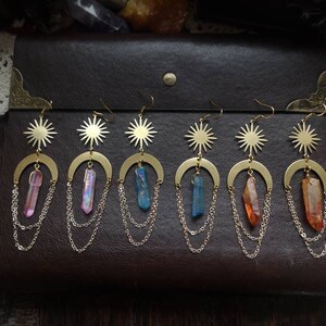 May include: Five pairs of gold earrings with a crescent moon, sun, and crystal pendant. The crystals are pink, blue, and orange.