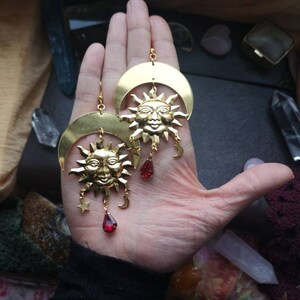 May include: Gold tone earrings with a crescent moon and sun design. Each earring features a red gemstone dangle.
