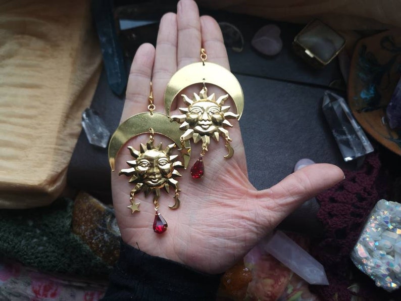 May include: Gold tone earrings with a crescent moon and sun design. Each earring features a red gemstone dangle.