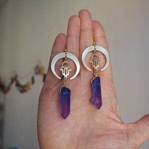 Puede incluir: Un par de pendientes de media luna plateados con un amuleto de hamsa dorado y un colgante de cristal morado.