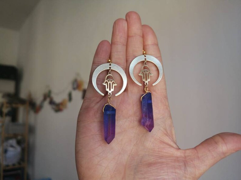 Puede incluir: Un par de pendientes de media luna plateados con un amuleto de hamsa dorado y un colgante de cristal morado.