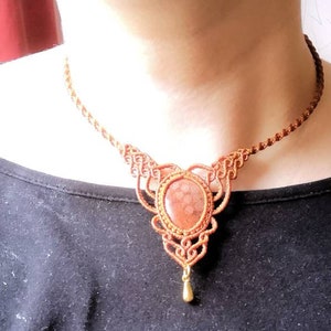 Puede incluir: Un collar de macram&eacute; marr&oacute;n con un colgante de piedra roja y marr&oacute;n. El colgante tiene forma de l&aacute;grima y tiene un acento de metal dorado.