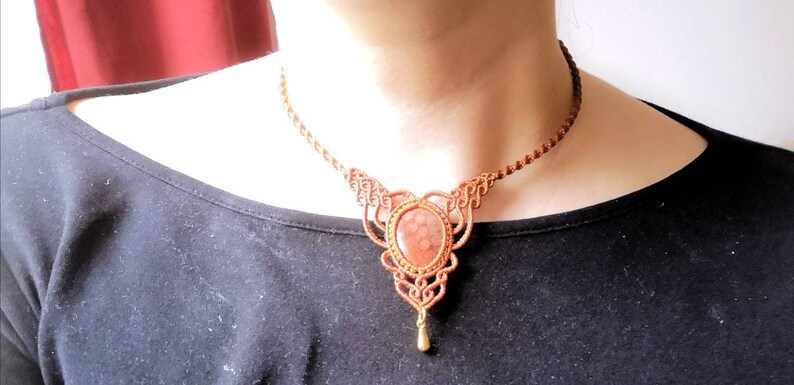 Puede incluir: Un collar de macram&eacute; marr&oacute;n con un colgante de piedra roja y marr&oacute;n. El colgante tiene forma de l&aacute;grima y tiene un acento de metal dorado.