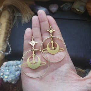 May include: Gold hoop earrings with a crescent moon and star design. The earrings are dangling from the hoops.