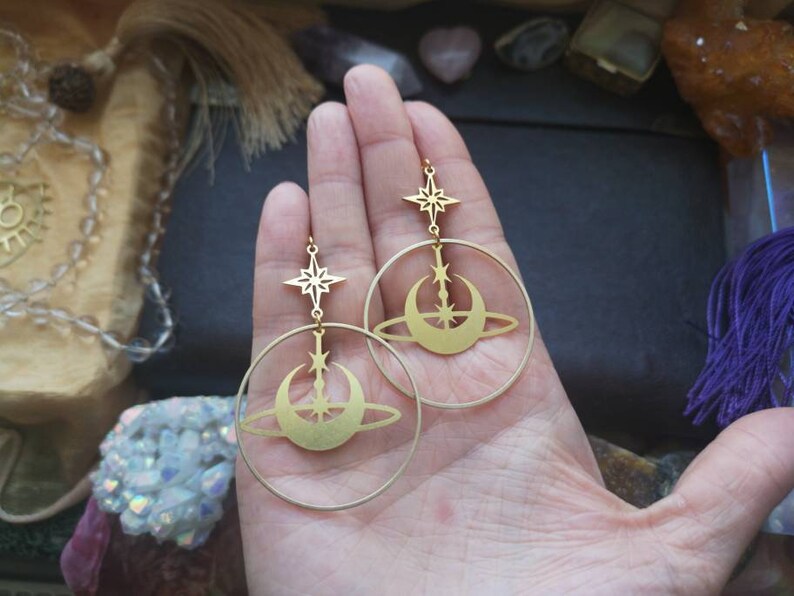 May include: Gold hoop earrings with a crescent moon and star design. The earrings are dangling from the hoops.