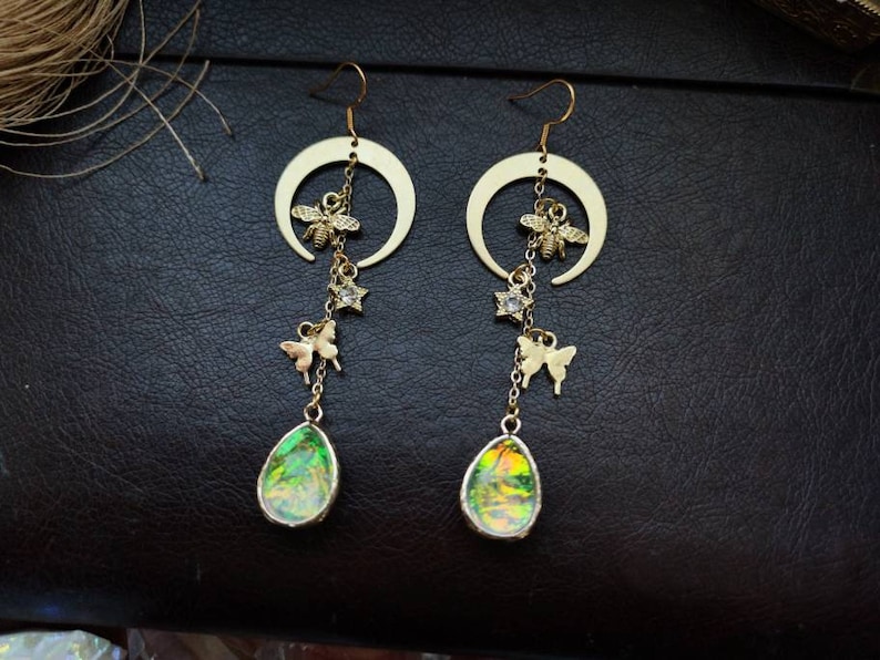 Puede incluir: Pendientes de media luna dorados con una piedra verde iridiscente en forma de l&aacute;grima colgante, un dije de abeja, un dije de mariposa y un dije de estrella.