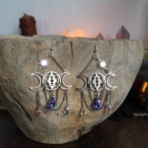 May include: Silver earrings with a triple moon, sun, and crystal accents. The earrings are dangling and have a delicate, ethereal look.