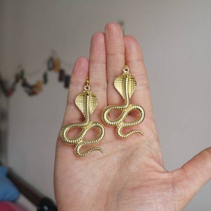 May include: A pair of gold-toned earrings featuring a detailed cobra snake design. The earrings are dangling and have a textured finish.