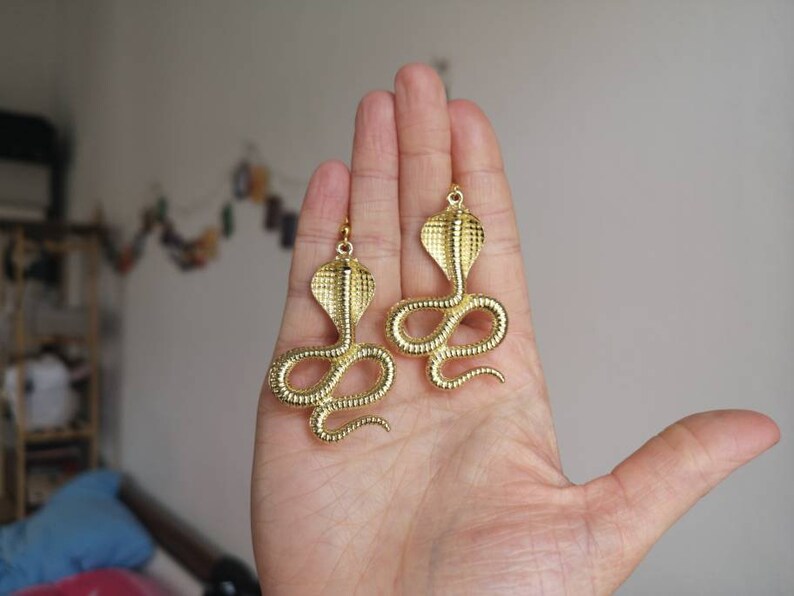May include: A pair of gold-toned earrings featuring a detailed cobra snake design. The earrings are dangling and have a textured finish.