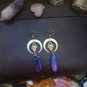Puede incluir: Un par de pendientes de oro con un dise&ntilde;o de media luna y un amuleto de mano de hamsa. Cada pendiente presenta un cristal morado y azul colgante.