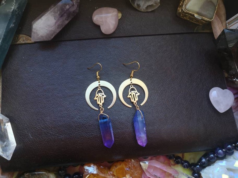Puede incluir: Un par de pendientes de oro con un dise&ntilde;o de media luna y un amuleto de mano de hamsa. Cada pendiente presenta un cristal morado y azul colgante.