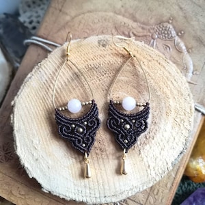 May include: A pair of brown macrame earrings with gold accents and a pink stone bead. The earrings are hanging from a wooden surface.