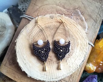 May include: A pair of brown macrame earrings with gold accents and a pink stone bead. The earrings are hanging from a wooden surface.