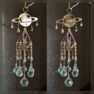 Könnte beinhalten: Ein goldfarbener Kristall-Sonnenfänger mit geometrischem Design und einem violetten Amethyst-Kristall, der in der Mitte hängt. Der Sonnenfänger hat einen Ring mit einer Sichelmond und Sternen und ist mit mehreren Kristalltropfen verziert.