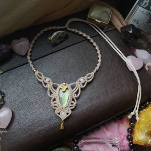 Puede incluir: Un collar de macram&eacute; blanco con un colgante de piedra preciosa verde y dorada. El collar se muestra en un diario de cuero marr&oacute;n con cristales rosas y morados.