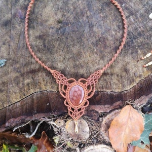 Puede incluir: Un collar de macram&eacute; marr&oacute;n con un colgante de piedra de cornalina roja. El collar est&aacute; hecho con un cord&oacute;n anudado y tiene un dise&ntilde;o simple.