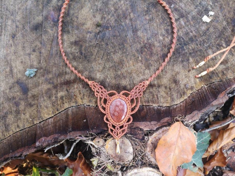 Puede incluir: Un collar de macram&eacute; marr&oacute;n con un colgante de piedra de cornalina roja. El collar est&aacute; hecho con un cord&oacute;n anudado y tiene un dise&ntilde;o simple.