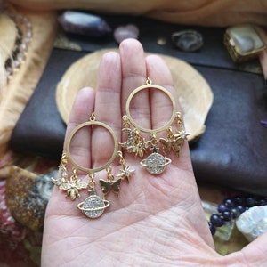 May include: Gold hoop earrings with dangling charms, including a bee, a butterfly, a crescent moon, a star, and a planet with a sparkly surface.