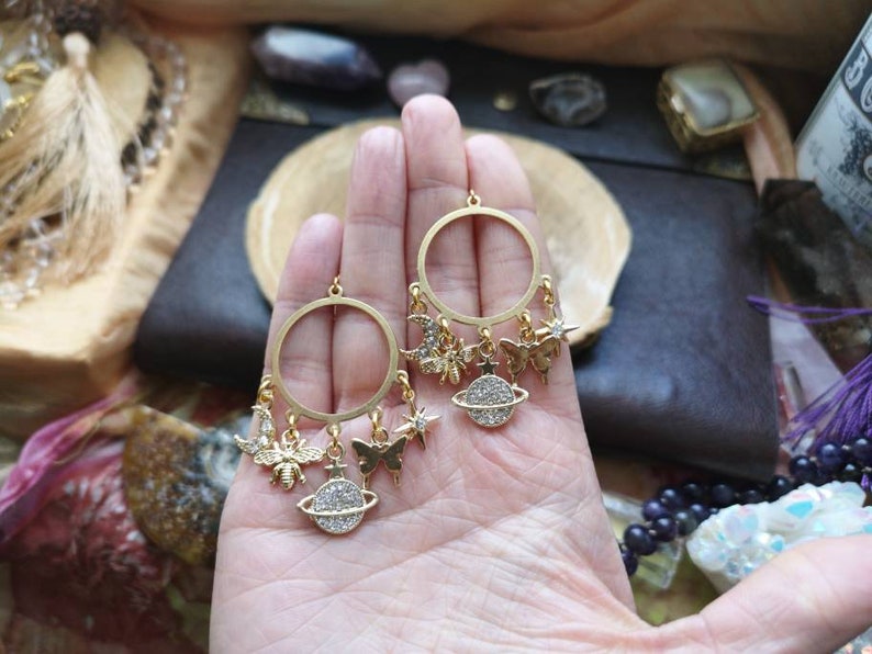 May include: Gold hoop earrings with dangling charms, including a bee, a butterfly, a crescent moon, a star, and a planet with a sparkly surface.