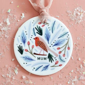 Può includere: Ornamento rotondo in ceramica bianca con un'illustrazione di pettirosso, foglie blu e bacche rosse. La parola "MUM" è stampata in nero. Un nastro rosa è attaccato per appenderlo. Lo sfondo è una superficie rosa chiaro con coriandoli iridescenti.