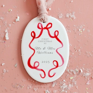 May include: White oval Christmas ornament with red ribbon design and text. The text reads "FIRST CHRISTMAS AS Mr & Mrs Williams 2025". A pink ribbon is attached for hanging. Festive holiday decor.