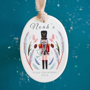May include: Oval Christmas ornament with a nutcracker design. The ornament is white with a red, black, and gold nutcracker illustration. The text "Noah's" and "First Christmas 2025" are also on the ornament. A beige ribbon is attached for hanging.