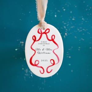 K&ouml;nnte beinhalten: Wei&szlig;er ovaler Weihnachtsschmuck mit rotem Schleifendesign und dem Text "FIRST CHRISTMAS AS Mr & Mrs Williams 2025". Der Schmuck wird von einem beigen Band vor einem t&uuml;rkisfarbenen Hintergrund gehalten.