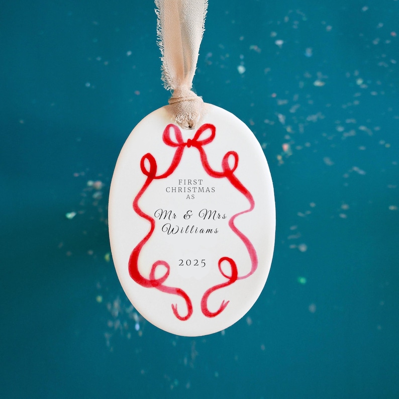 K&ouml;nnte beinhalten: Wei&szlig;er ovaler Weihnachtsschmuck mit rotem Schleifendesign und dem Text "FIRST CHRISTMAS AS Mr & Mrs Williams 2025". Der Schmuck wird von einem beigen Band vor einem t&uuml;rkisfarbenen Hintergrund gehalten.