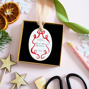 K&ouml;nnte beinhalten: Ein wei&szlig;er, ovaler Weihnachtsschmuck mit roten Banddetails und dem Text "First Christmas As Mr & Mrs Williams 2025" wird in einer schwarzen Box mit goldenem Innenraum pr&auml;sentiert. Der Schmuck h&auml;ngt an einem beigen Band. Der Hintergrund ist rosa mit dekorativen Elementen.