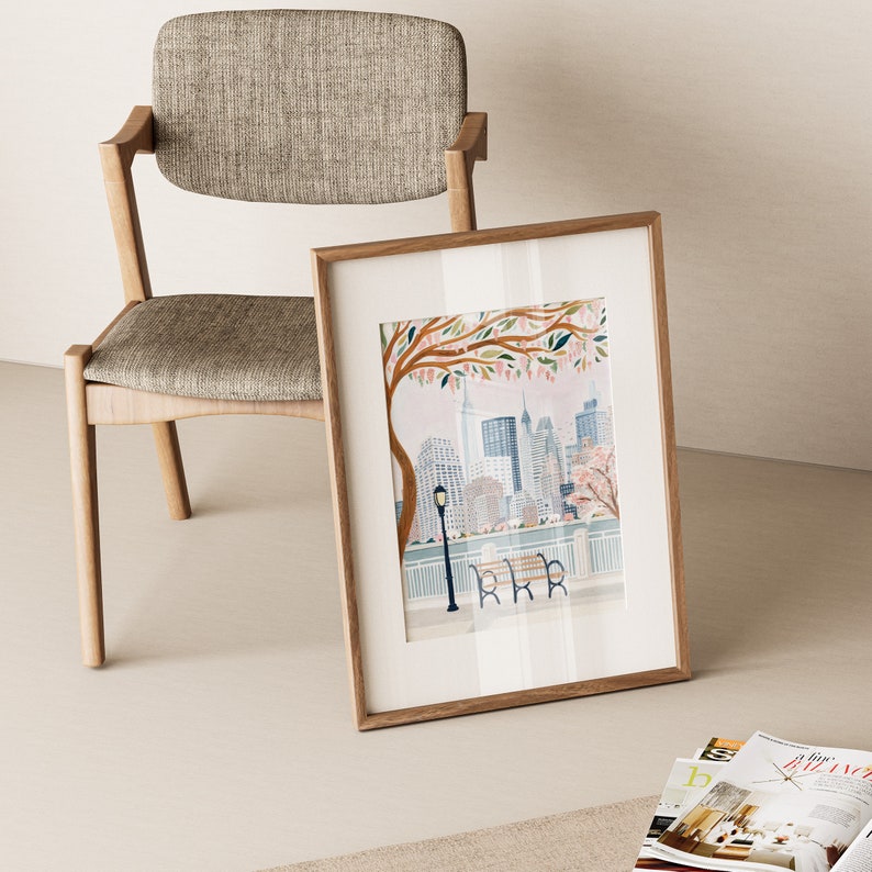 May include: A framed print of a cityscape with a pink and white flowering tree in the foreground. The city is depicted in a stylized, whimsical style with buildings in shades of blue and gray. The print is in a light wood frame.