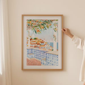 May include: A framed watercolor print depicting a European cityscape with orange trees in the foreground. The artwork features buildings with red tile roofs, blue and white patterned tiles, and a view of the sea. The frame is made of light-colored wood.