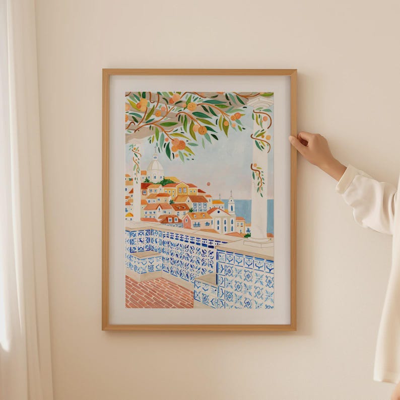 May include: A framed watercolor print depicting a European cityscape with orange trees in the foreground. The artwork features buildings with red tile roofs, blue and white patterned tiles, and a view of the sea. The frame is made of light-colored wood.