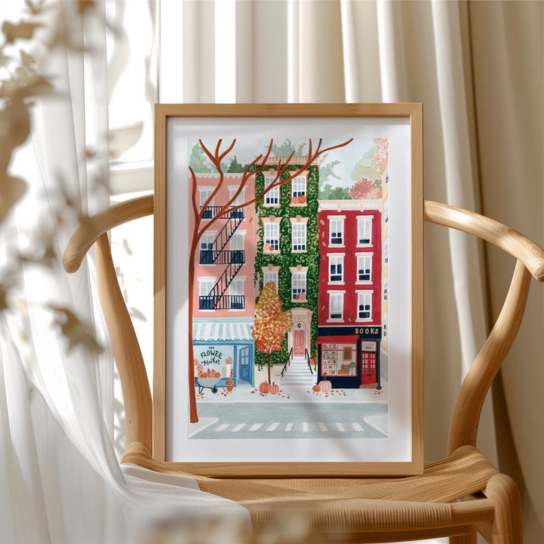 May include: A framed art print showing a street scene with vibrant buildings. The buildings are pink, green, and red, with a flower market and a bookstore. The print is displayed on a wooden chair.